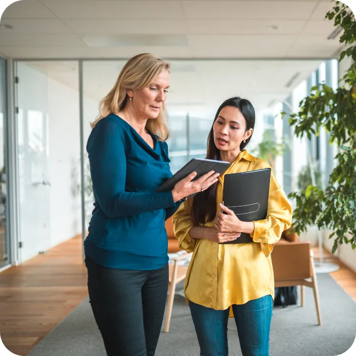 Two women discussing documents in a bright office hallway.