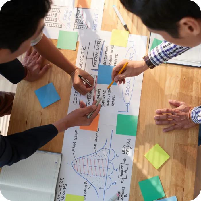 Three people collaborate on a diagram with sticky notes on a wooden table.