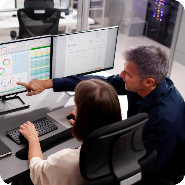 Man and woman in office analyzing data on dual monitors.