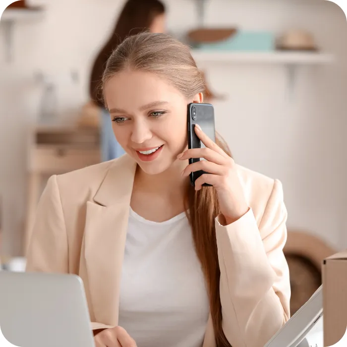Smiling woman in beige blazer on phone, working at a laptop.
