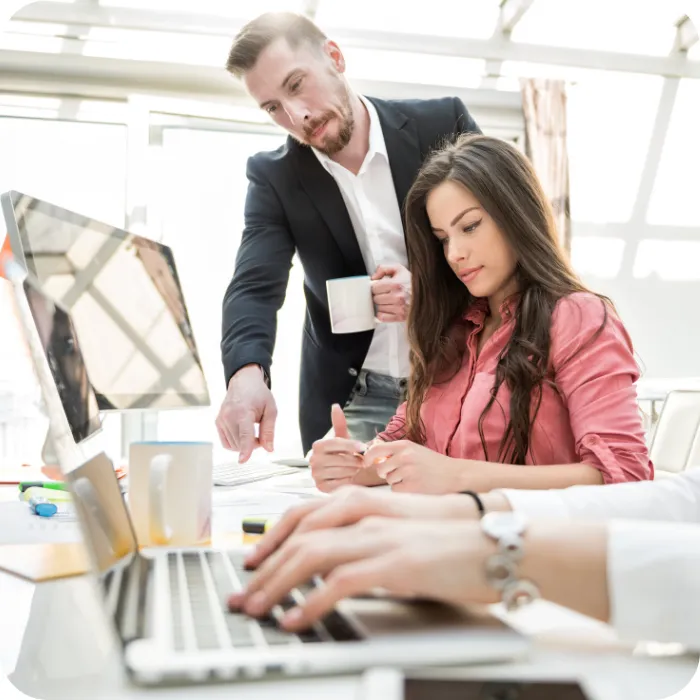 Man with coffee cup discussing work with woman at a desk with a laptop.