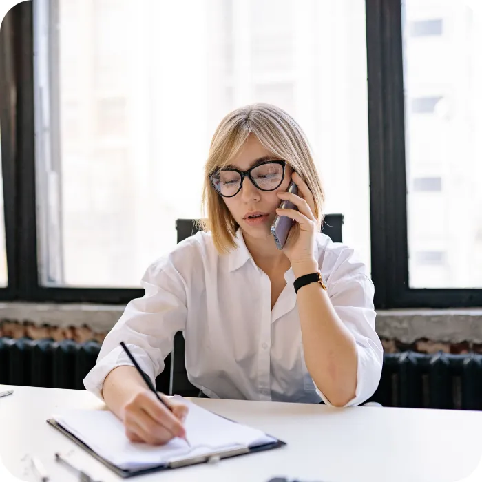 Woman in a white shirt, talking on phone, writing in a notebook.