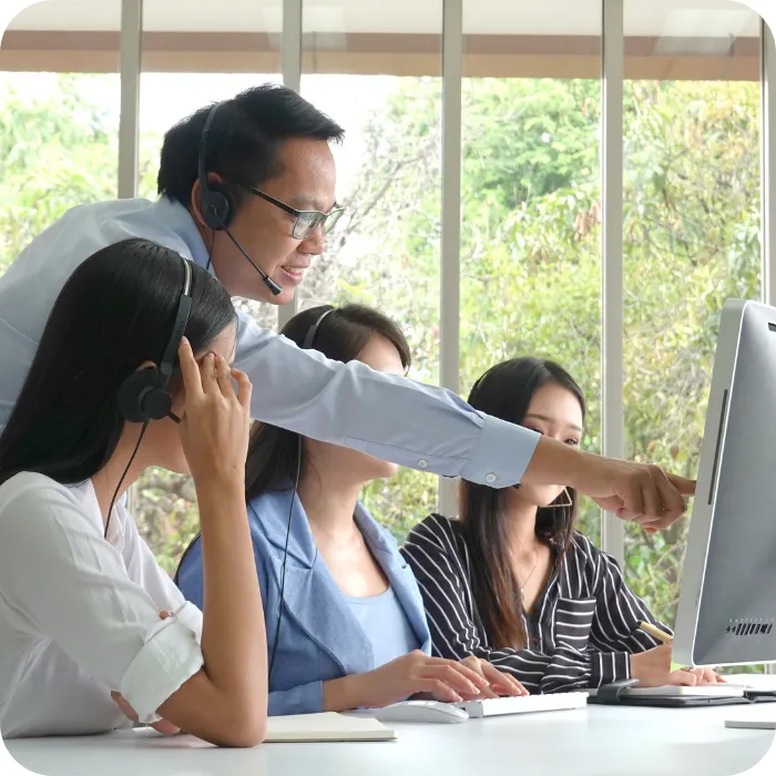 Man in headset pointing at computer screen, guiding three seated women.
