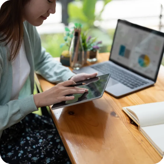 Person using a tablet at a desk with a laptop and notebook.