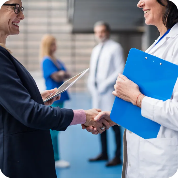 Two professionals shaking hands in a medical setting, one holding a blue clipboard.