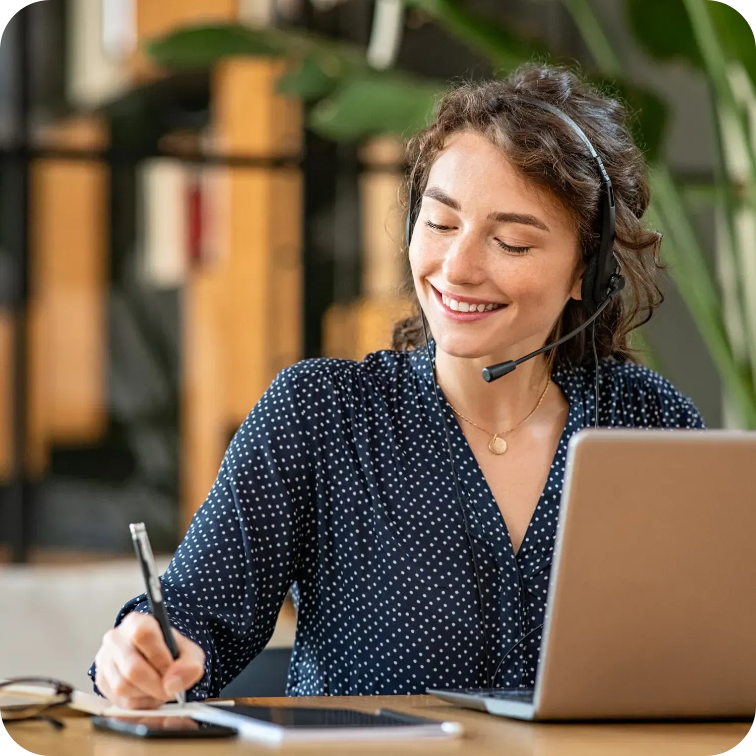 Smiling woman with headset writing, laptop open, in office setting.