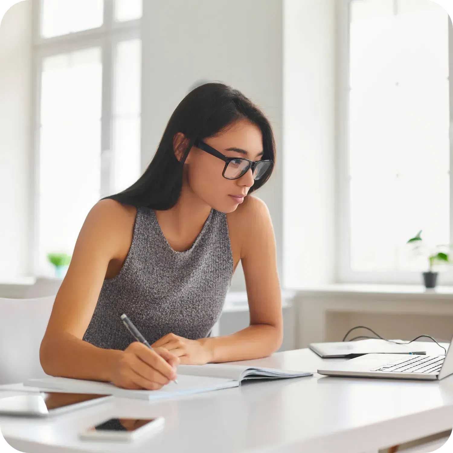 Woman wearing glasses, writing at a desk with a laptop.