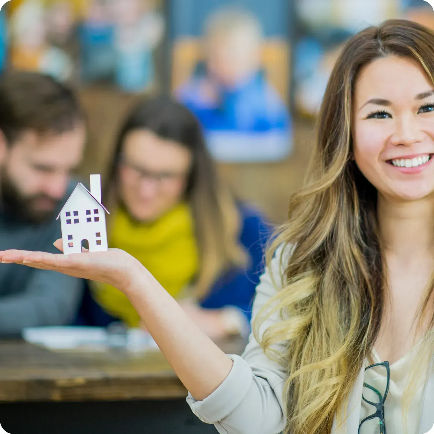 Woman smiling, holding a small house model, with blurred people in the background.