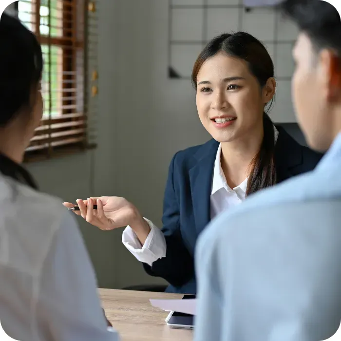 A woman in a blazer smiles while speaking to two people in an office setting.
