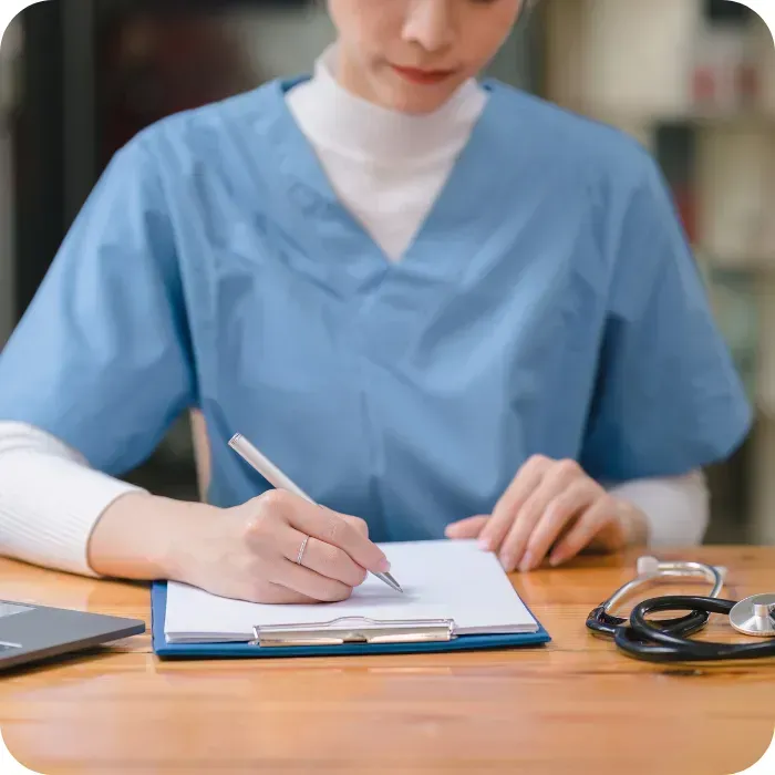 Nurse writing on clipboard at desk, stethoscope nearby.