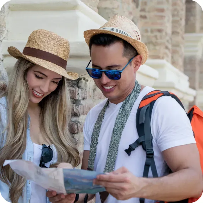 Tourists with hats and backpacks reading a map, smiling in a sunny setting.