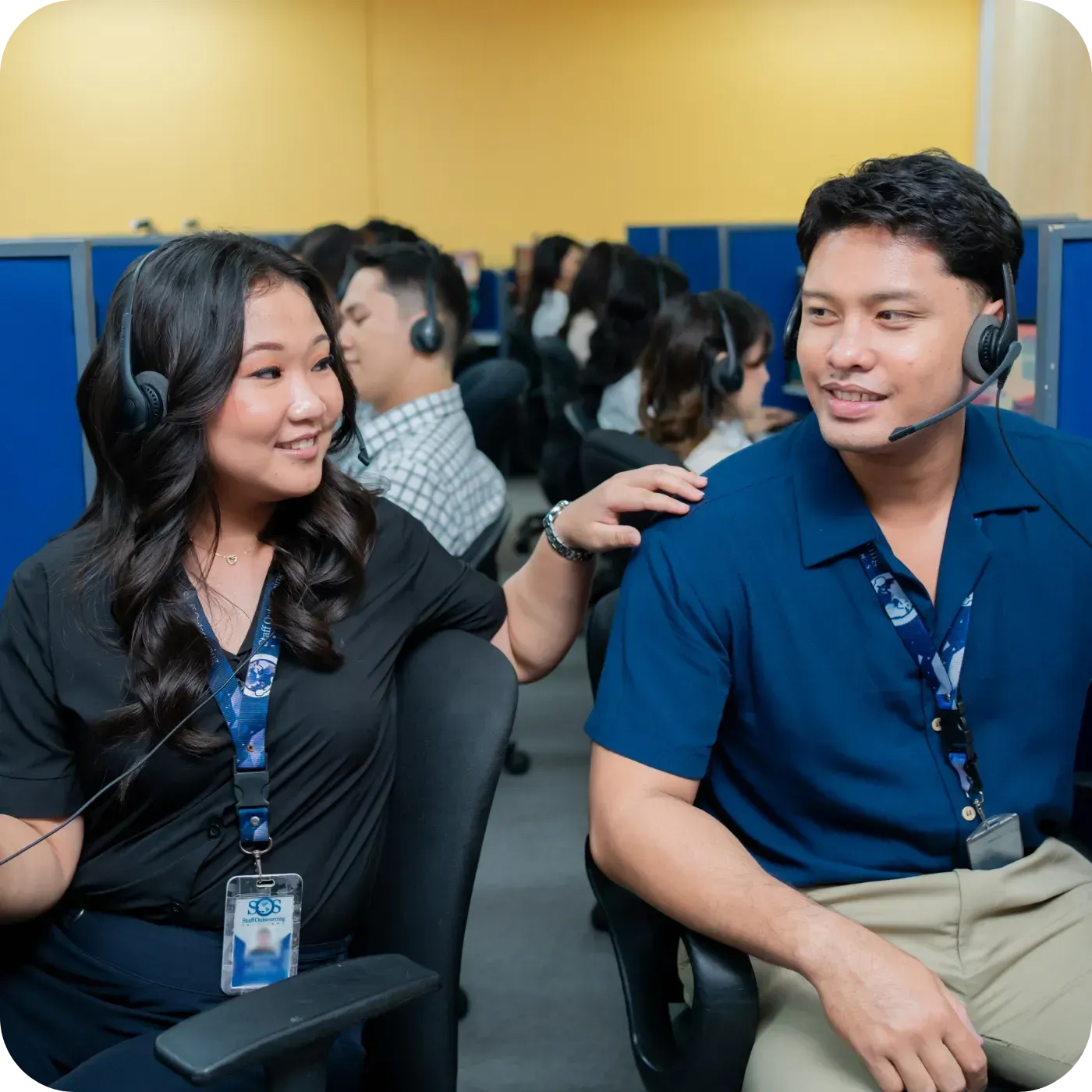 Two people smiling with headsets in a busy office.