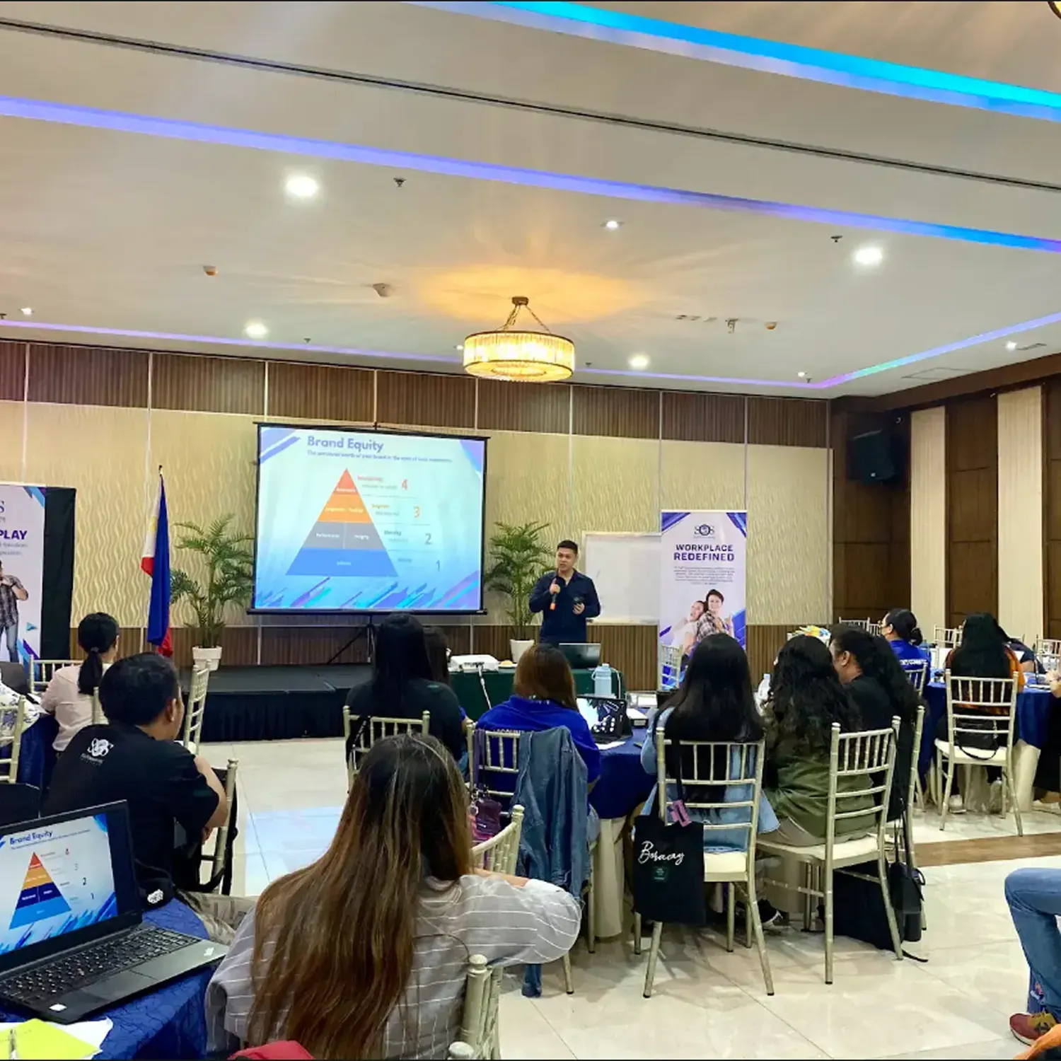 Conference room with a speaker presenting a pyramid chart to seated attendees.