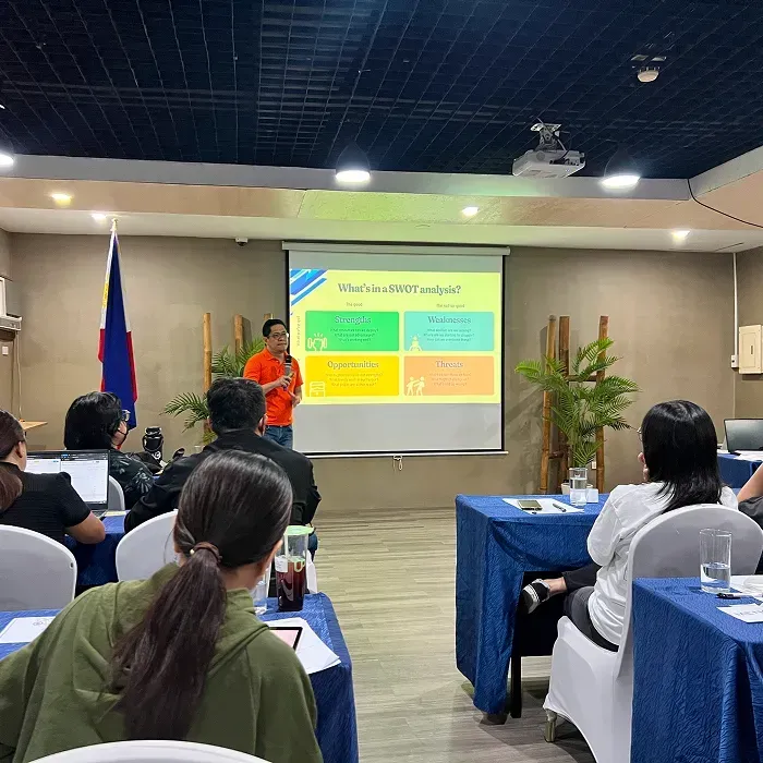 Conference room with a presenter in orange shirt speaking to seated attendees.