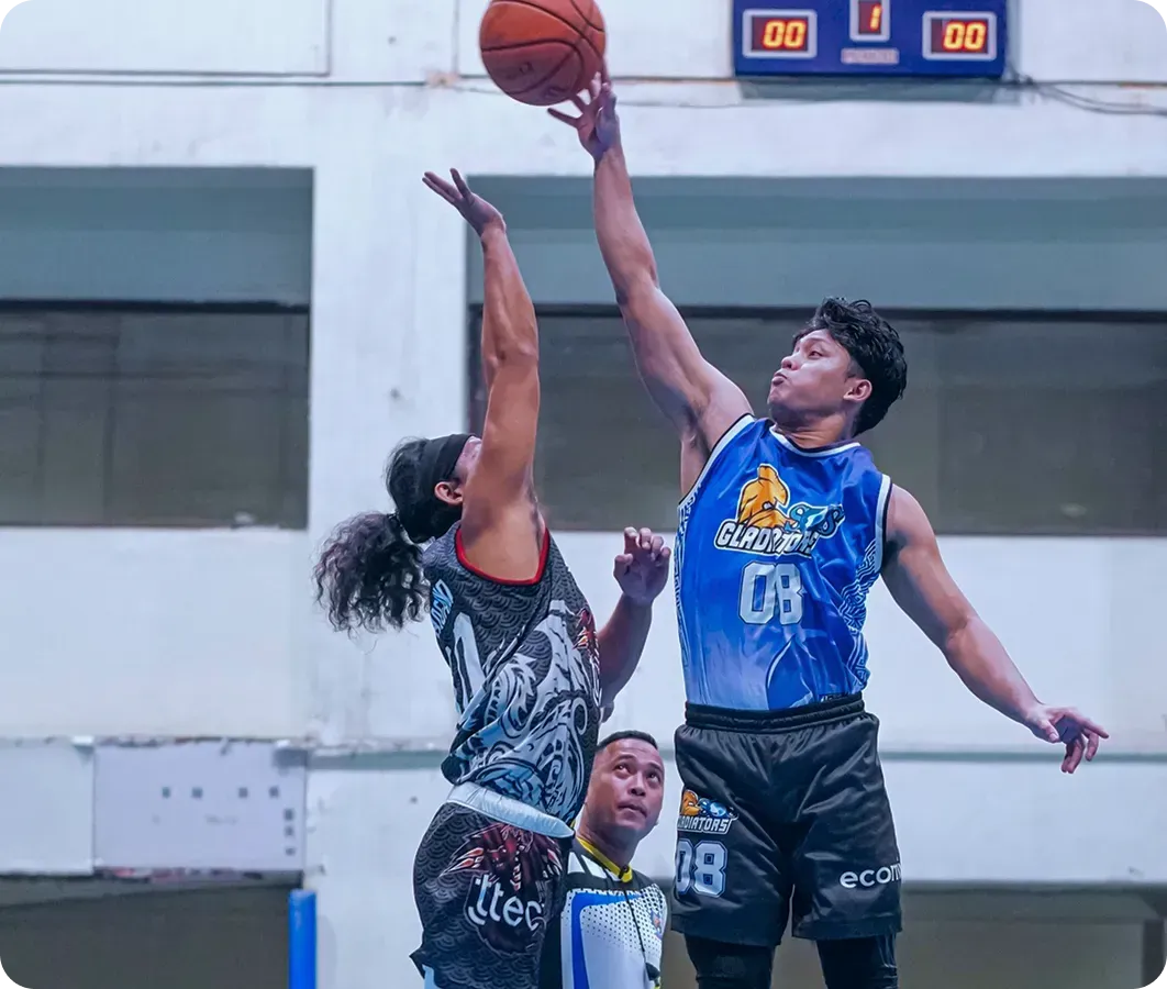 Two basketball players jump for the ball in an indoor court, with a scoreboard above.