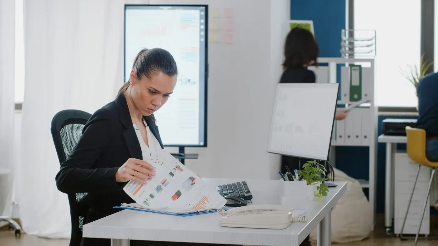 Office worker reviewing data on screen against printed reports at a desk